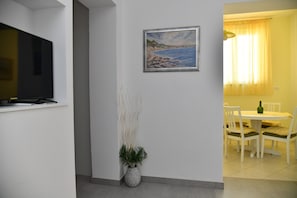 Une pièce dotée d’un téléviseur à écran plat fixé au mur, d’un tableau représentant une scène de plage, d’un vase contenant des plantes séchées et d’un coin repas avec une table et des chaises.