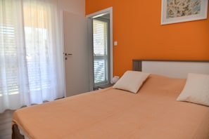 Une chambre avec un mur orange, un lit avec des oreillers blancs, une porte avec un miroir et une fenêtre avec des stores.