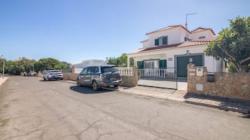 Una calle residencial con coches estacionados, un acceso con rejas y una casa con techo de tejas.