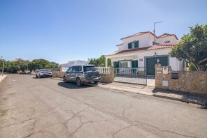 A residential street with parked cars, a gated entrance, and a house with a tiled roof.
