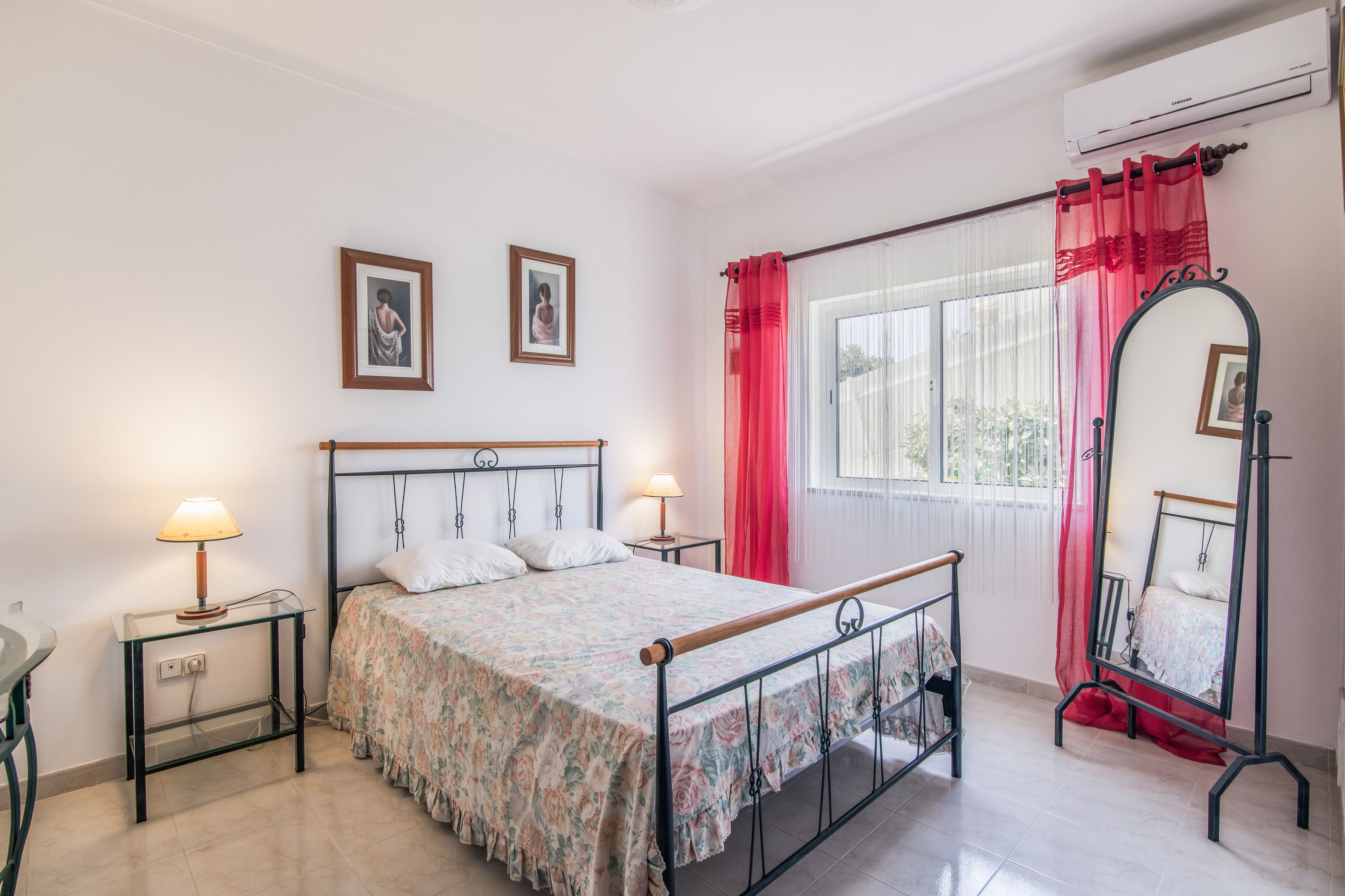 A bedroom with a bed, two bedside tables, a mirror, and a window with curtains.