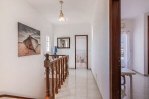 A hallway with a wooden staircase, a painting of a boat, a blue lamp, and a mirror reflecting a bathroom.