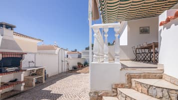 Un patio con suelo de piedra, una parrilla de barbacoa y una escalera blanca que conduce a una zona cubierta.