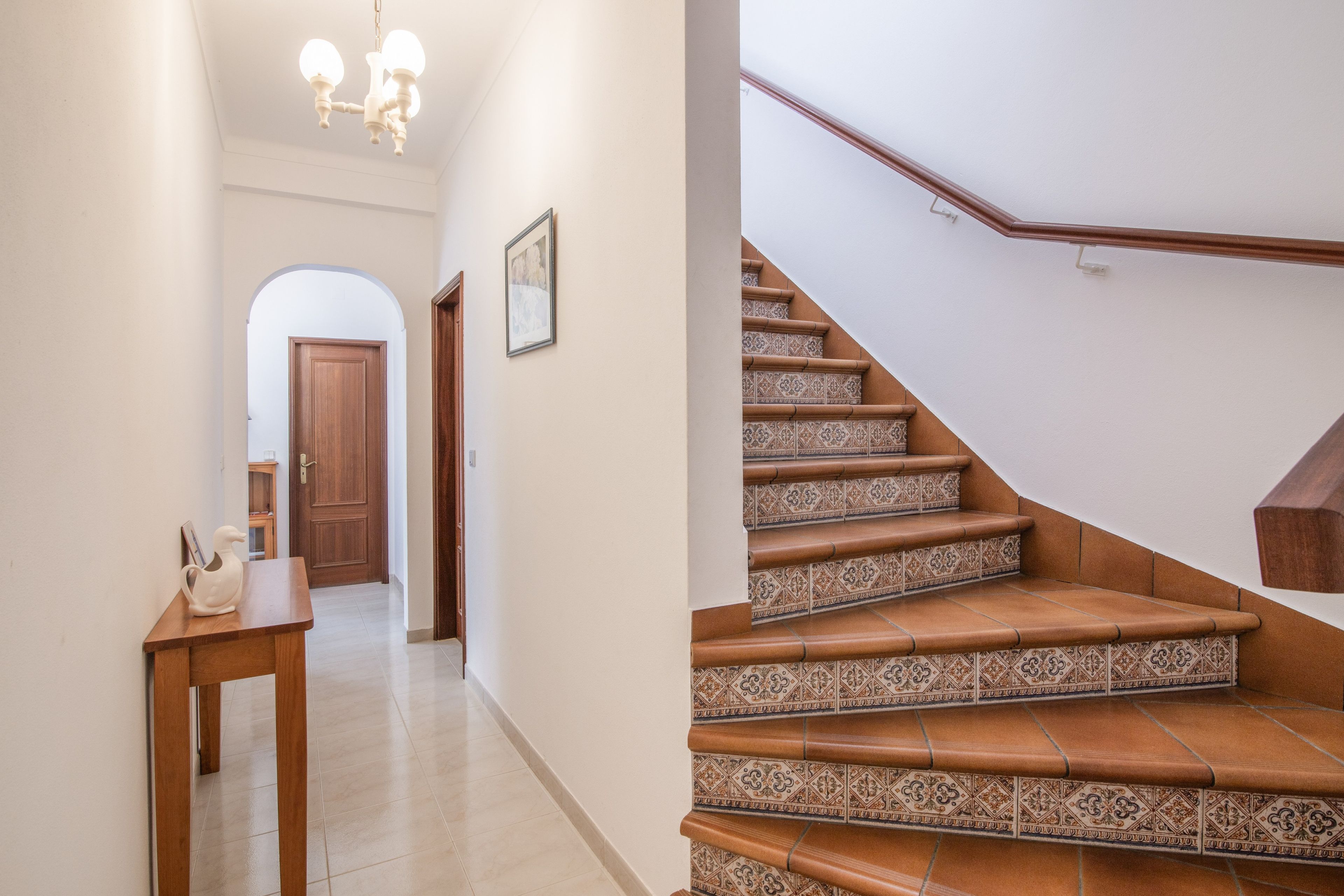 Un couloir avec une porte en bois, une petite table ornée d’un objet décoratif et un escalier dont les marches sont recouvertes de carreaux à motifs.