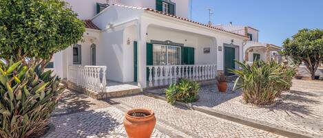 A white building with green shutters, a tiled roof, and a paved courtyard with potted plants.