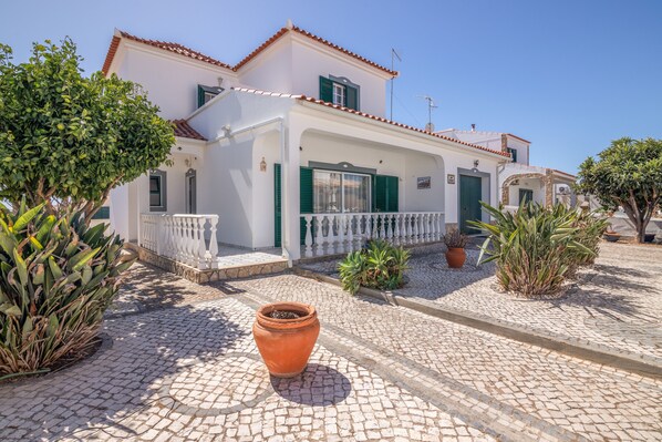 A white building with green shutters, a tiled roof, and a paved courtyard with potted plants.
