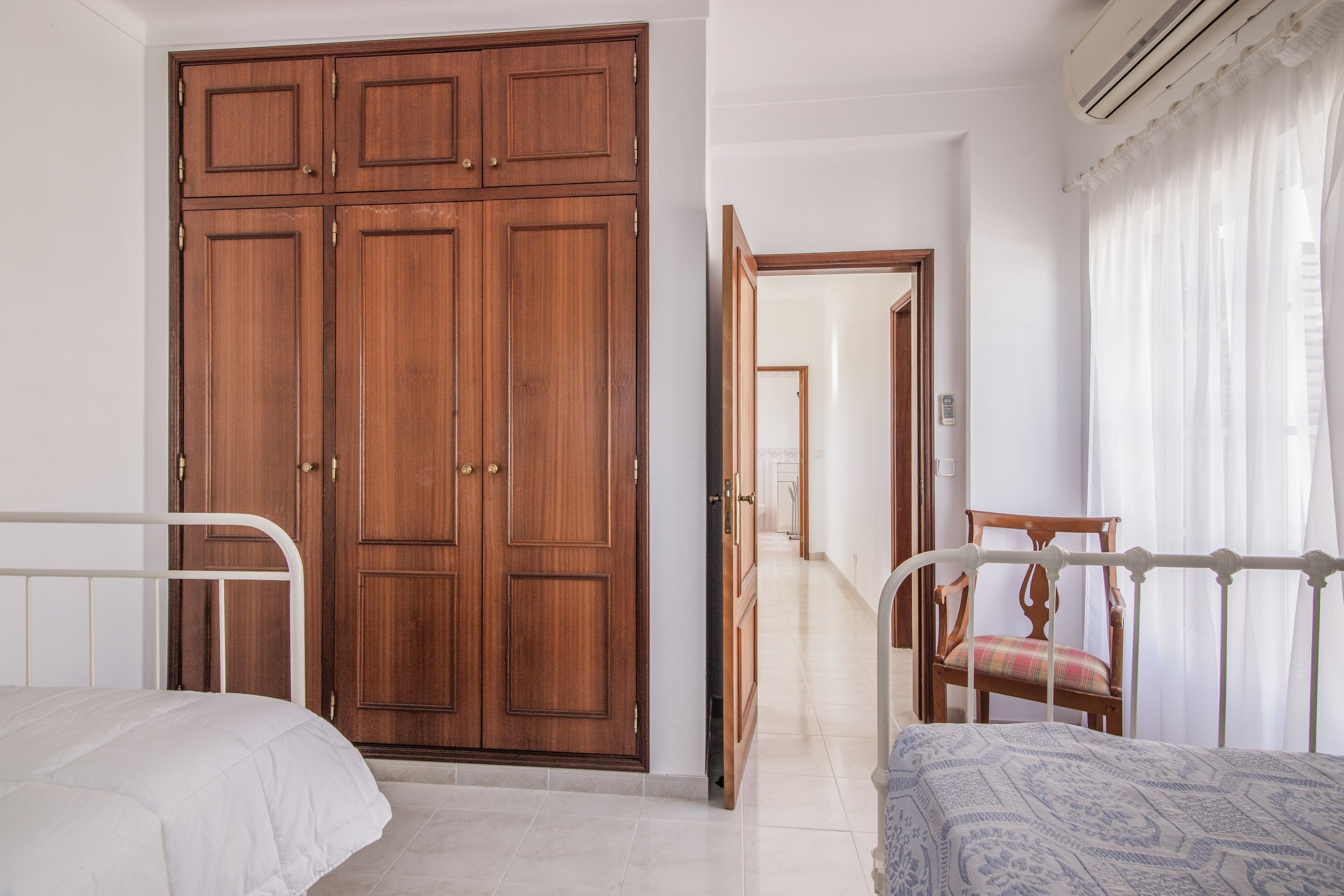 Un dormitorio con un armario de madera grande, una cama con colchón blanco y una silla con cojín estampado.