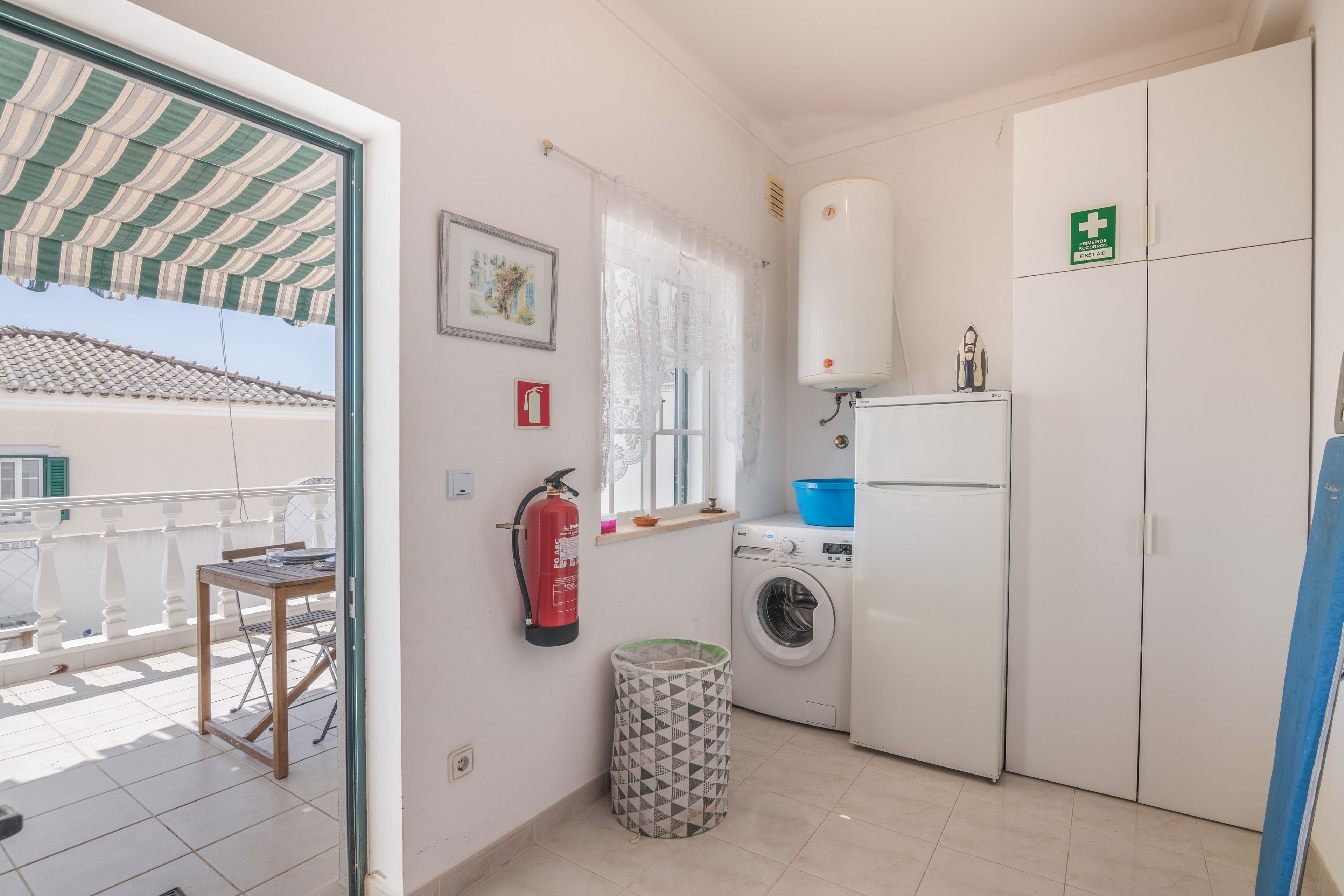 Een kleine keuken met een wasmachine, een brandblusser en een bord met daarop een eerste hulp symbool.