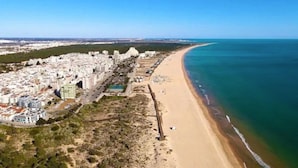 Una vista aerea di una città costiera con una lunga spiaggia, una zona urbana densa e cielo sereno.