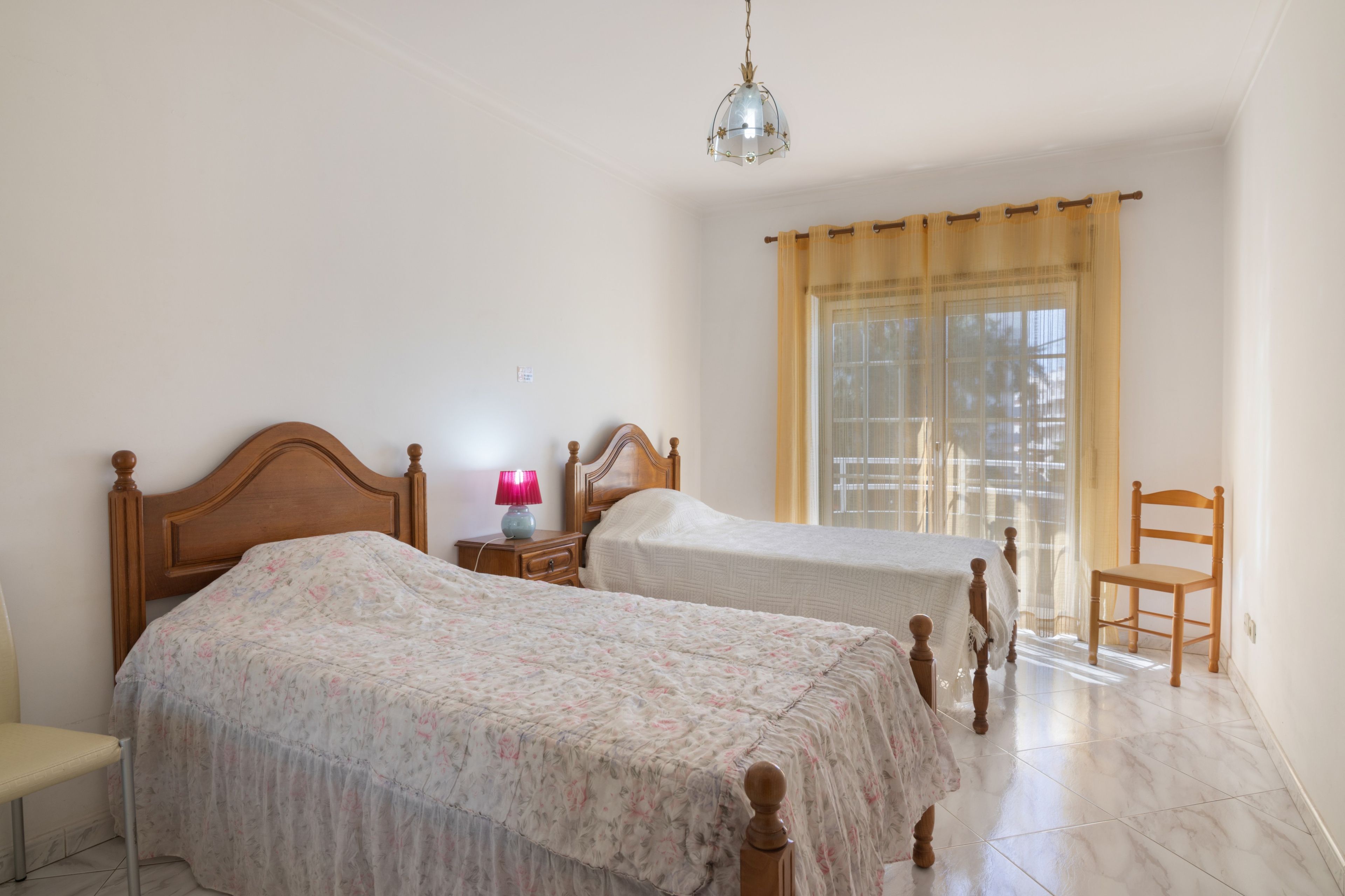 Un dormitorio con dos camas de madera, una mesita de noche con lámpara, una silla y una ventana con cortinas.