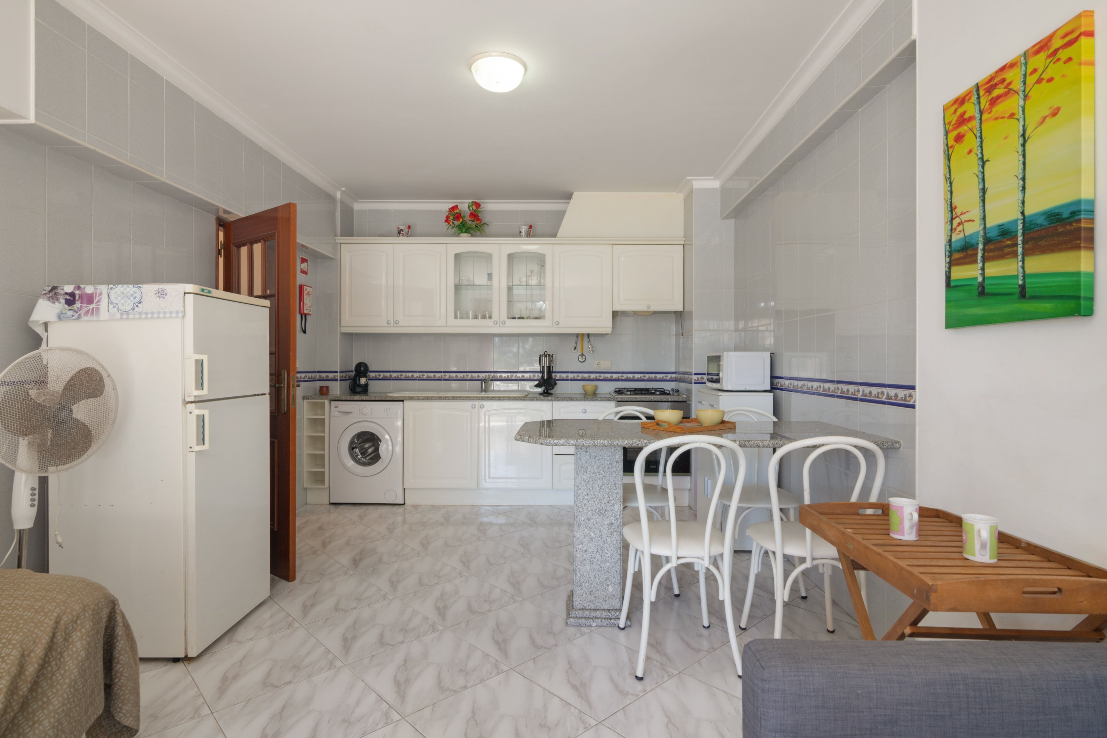 Une cuisine compacte avec des meubles blancs, un lave-linge, un coin repas avec une table et des chaises, et un ventilateur.