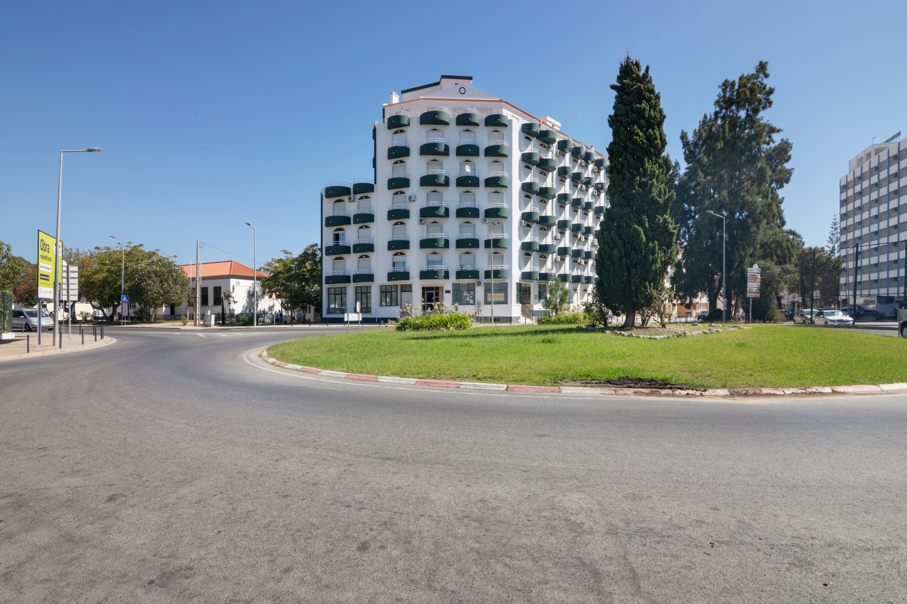 Un immeuble à plusieurs étages, doté d’une façade blanche et de balcons verdoyants, situé sur un rond-point avec un terre-plein central recouvert de gazon.