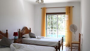 Una camera d'albergo con due letti, un comodino con una lampada, una sedia, un ventilatore e una finestra con tende.