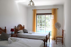Ett hotellrum med två sängar, ett sängbord med en lampa, en stol, en bordsfläkt och ett fönster med gardiner.