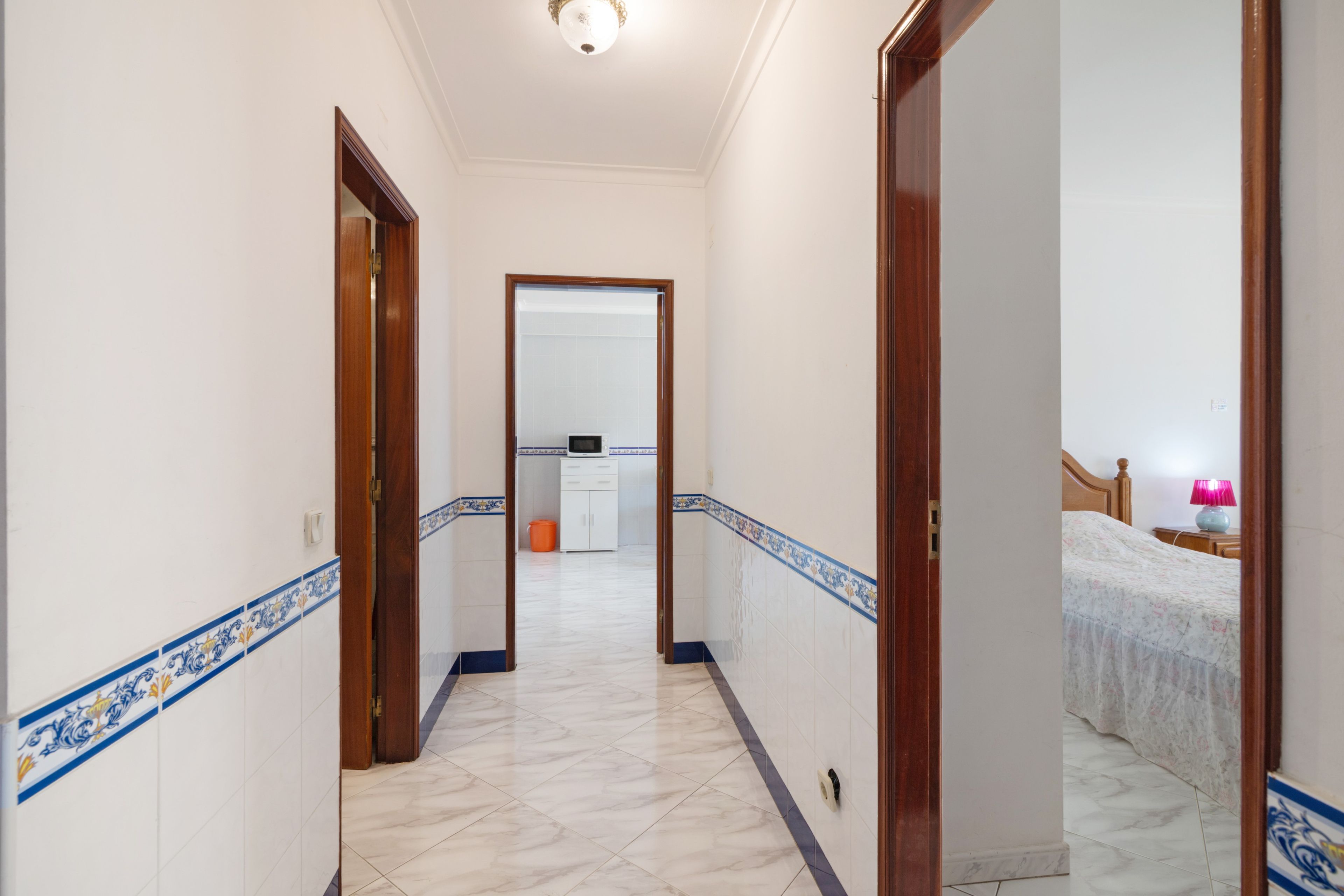 Un couloir aux murs blancs, orné de motifs décoratifs bleus, et dont le sol est recouvert de carreaux, mène à une chambre à coucher équipée d’un lit et d’une table de chevet.