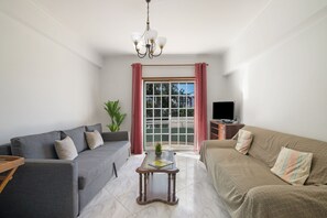 Un soggiorno con un divano grigio, un divano marrone, un tavolino in vetro, una TV su un mobile in legno e una finestra con tende rosa.