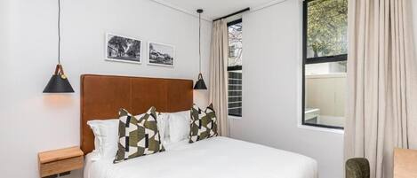 Quarto com uma cama grande, mesa de cabeceira de madeira, uma cadeira verde e janela com cortinas.