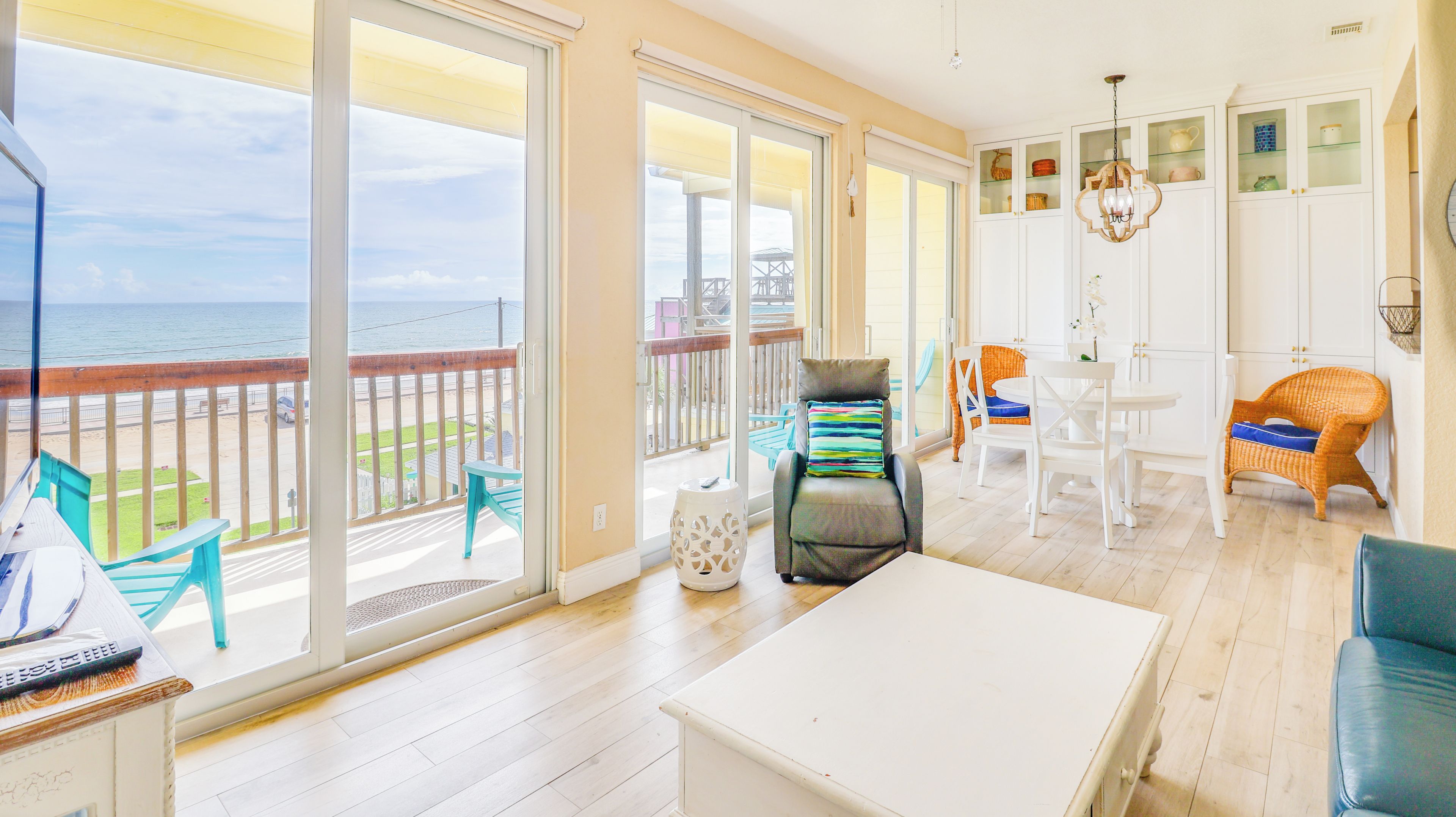 A spacious balcony with a view of the ocean, a dining area with white furniture, and a kitchen with white cabinets and a hanging light fixture.
