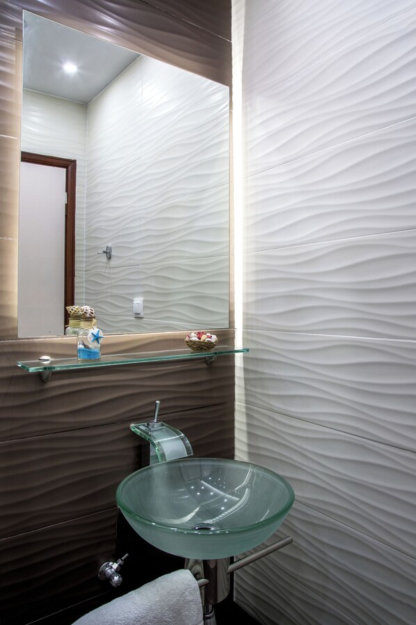 A bathroom with a glass sink, a mirror, and a shelf with decorative items.