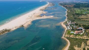 Oversigtsbillede af et kystområde med en sandstrand, klart blåt vand og et boligområde med huse og grønne områder.