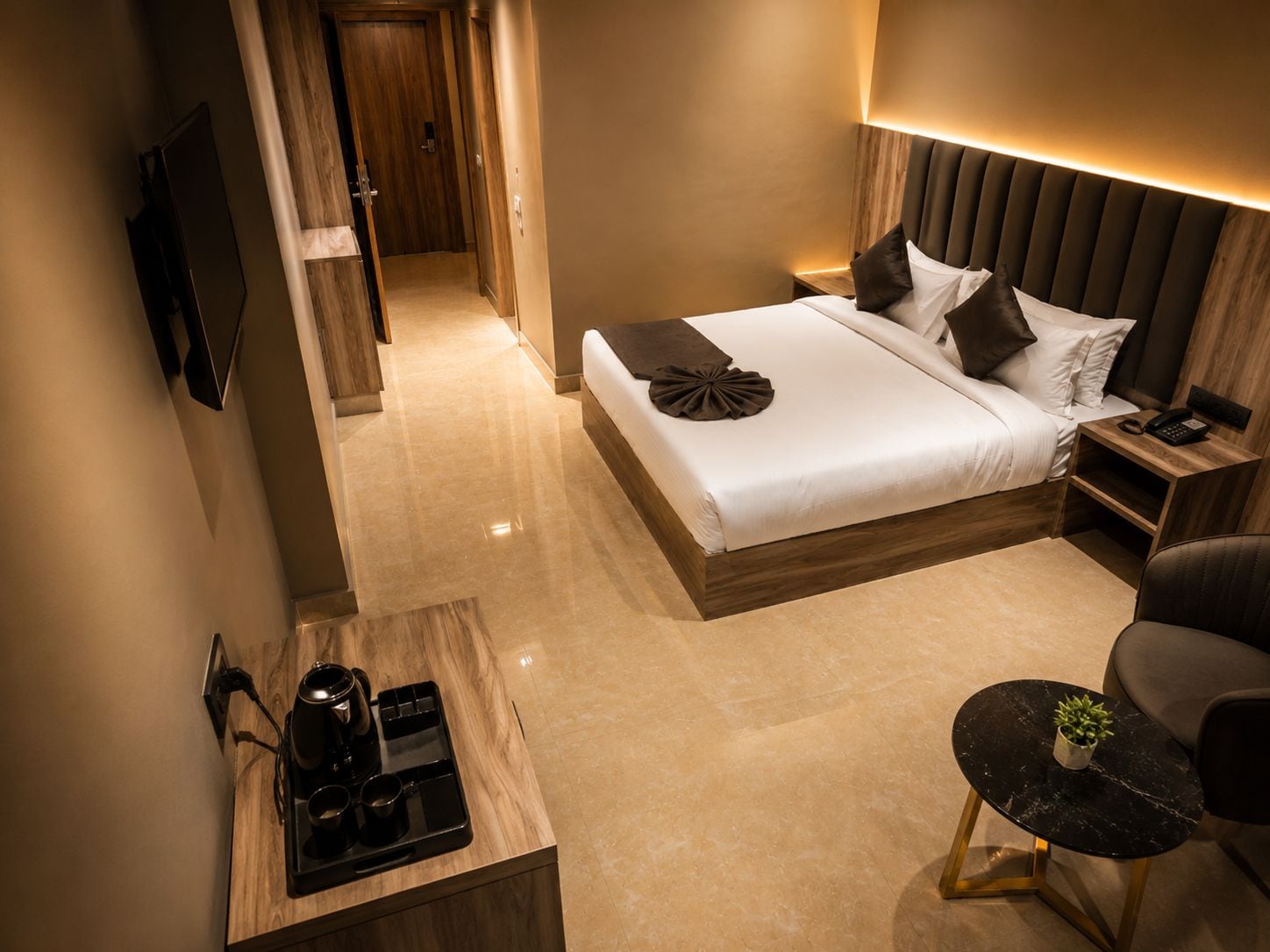 Una habitación de hotel moderna con una cama grande, mesitas de noche, un escritorio con cafetera, una silla y una pequeña mesa con una planta.