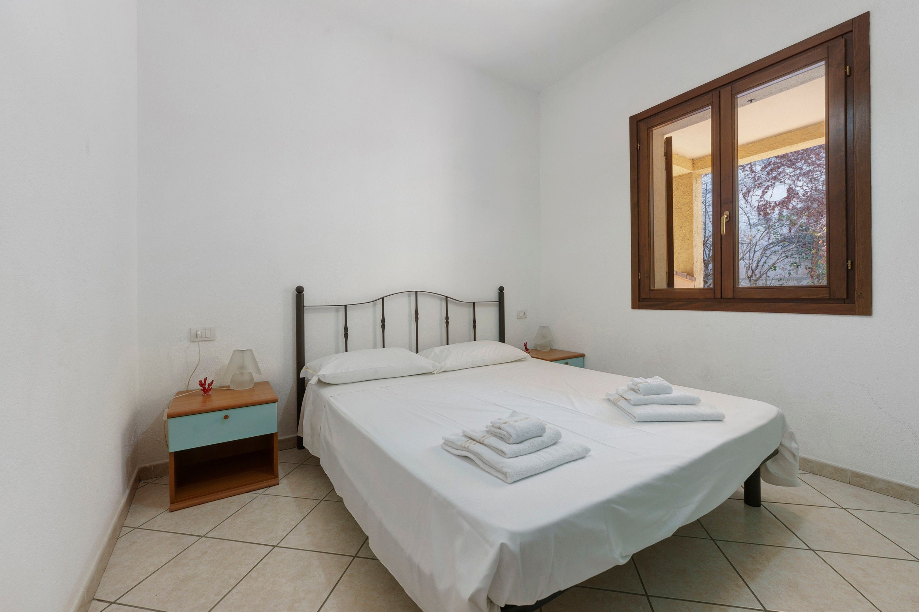 Un lit simple avec du linge de lit et des serviettes blanches, une table de chevet avec une lampe et une fenêtre donnant sur des arbres.