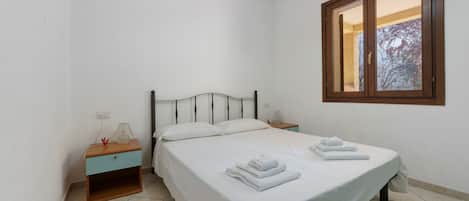 Un lit simple avec du linge de lit et des serviettes blanches, une table de chevet avec une lampe et une fenêtre donnant sur des arbres.