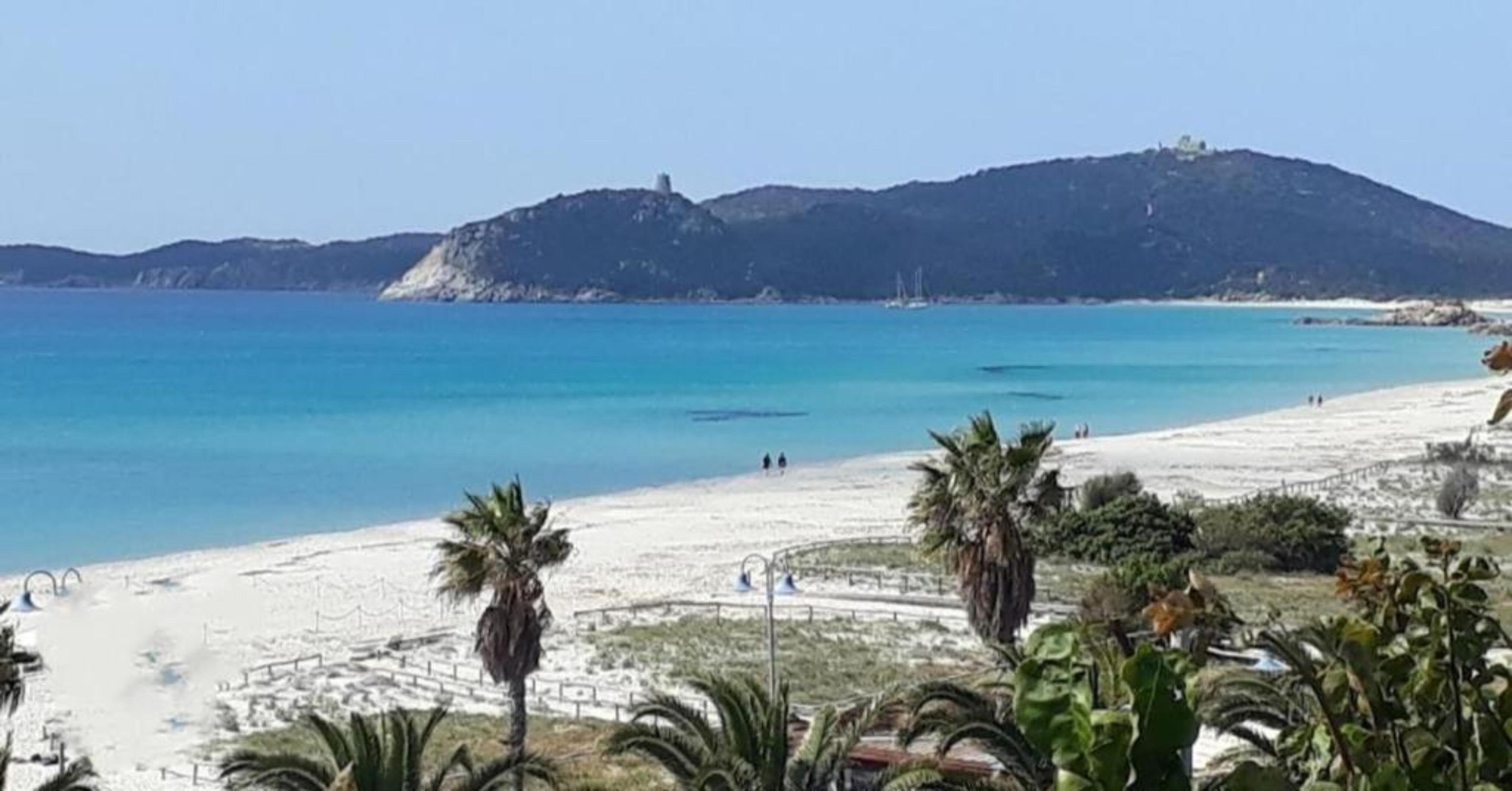 A beach with clear blue water, a sandy shore, and a hill in the background.