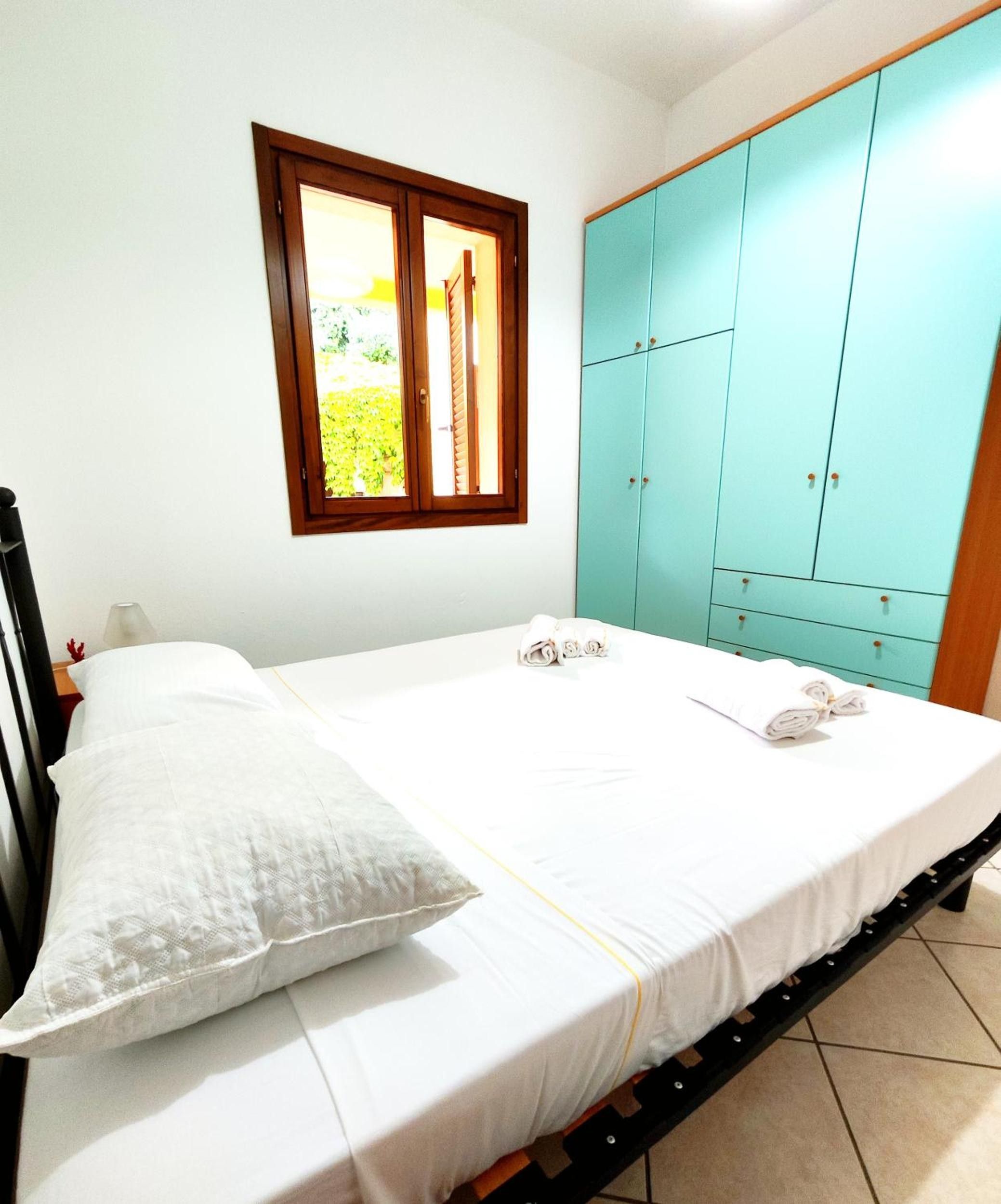 A single bed with a white mattress and a patterned pillow, a wooden headboard, a window with a view of greenery, and a teal-colored wardrobe with multiple drawers.