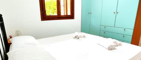 A single bed with a white mattress and a patterned pillow, a wooden headboard, a window with a view of greenery, and a teal-colored wardrobe with multiple drawers.