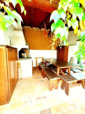 Uma cozinha rústica ao ar livre com parede de tijolos, bancos de madeira e forno de pedra.