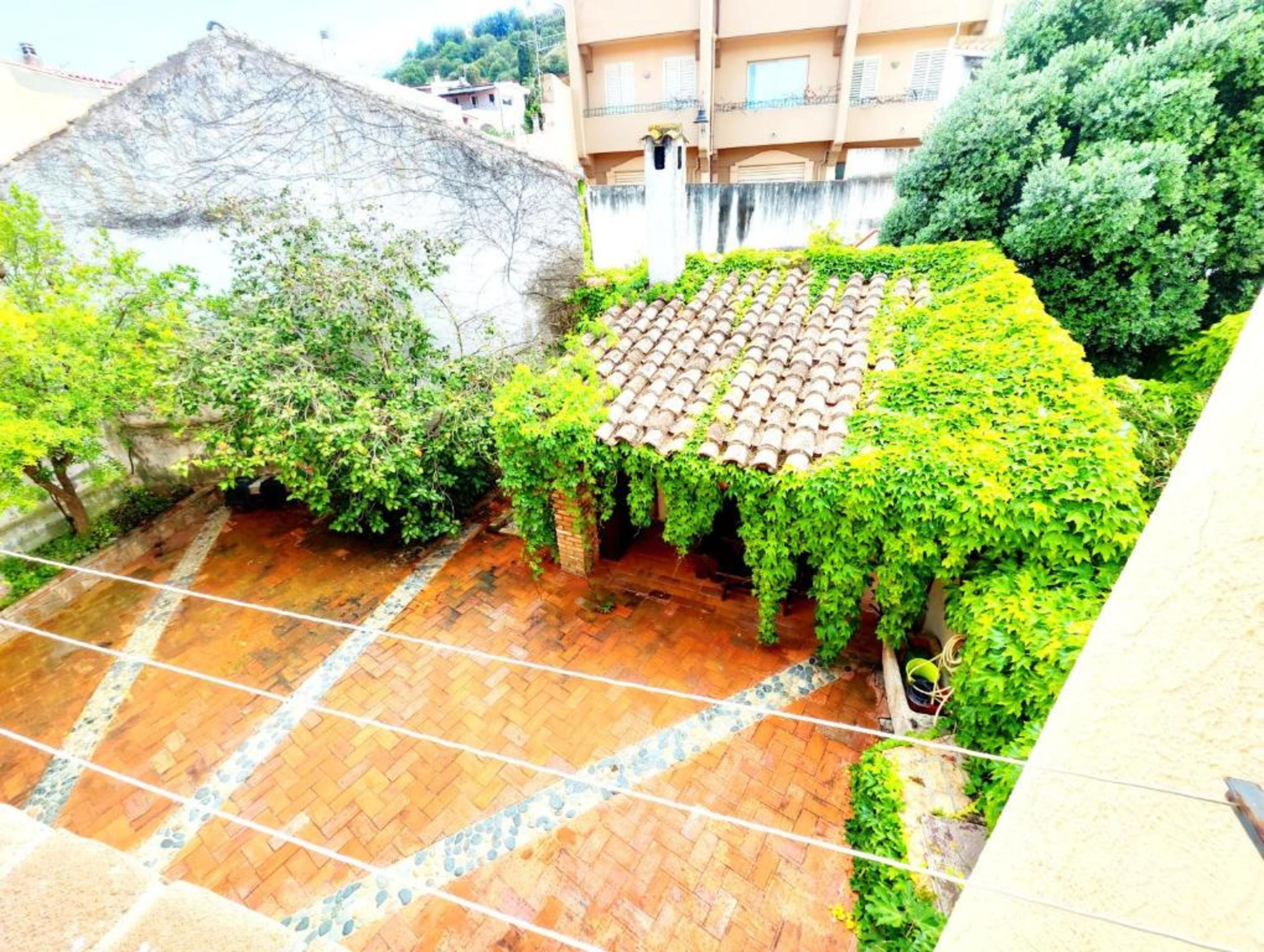 A courtyard with a tiled floor, surrounded by greenery and a building with a tiled roof.