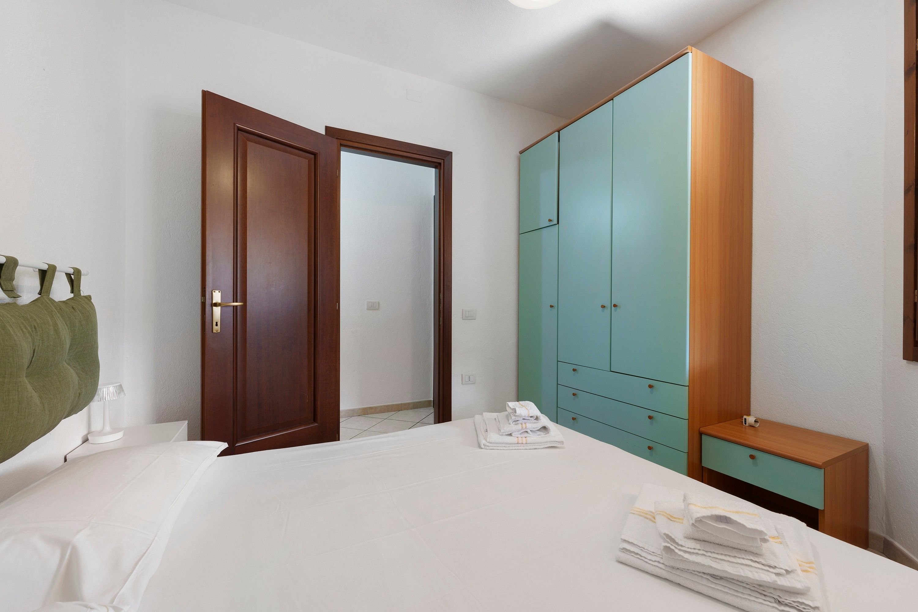 A hotel room with a bed, a wardrobe, a bedside table, and a green towel.