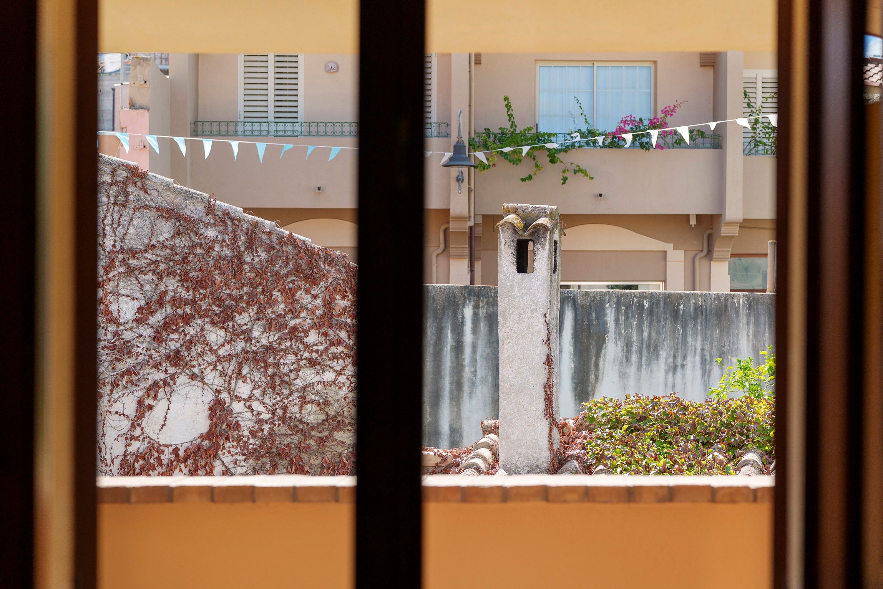 A view through a window with a stone wall, a metal fence, and a building with balconies and windows in the background.