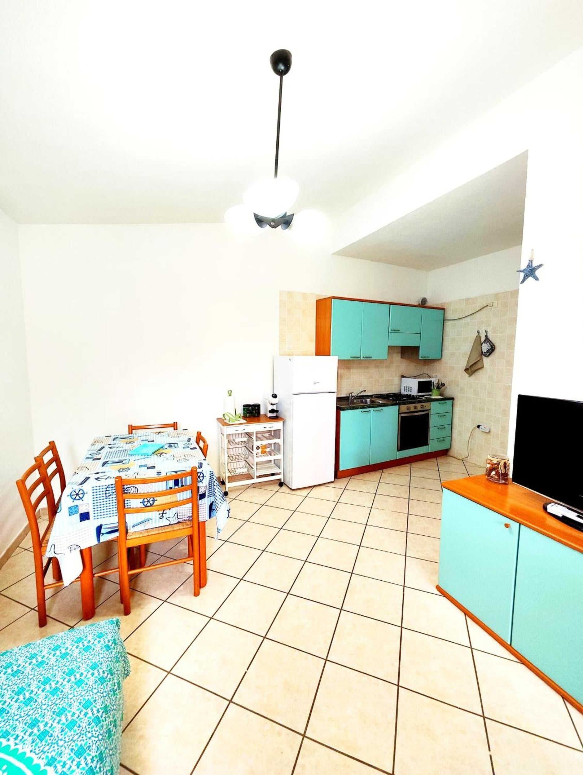 A compact kitchen with teal cabinets, a white refrigerator, and a dining area with a table and chairs.