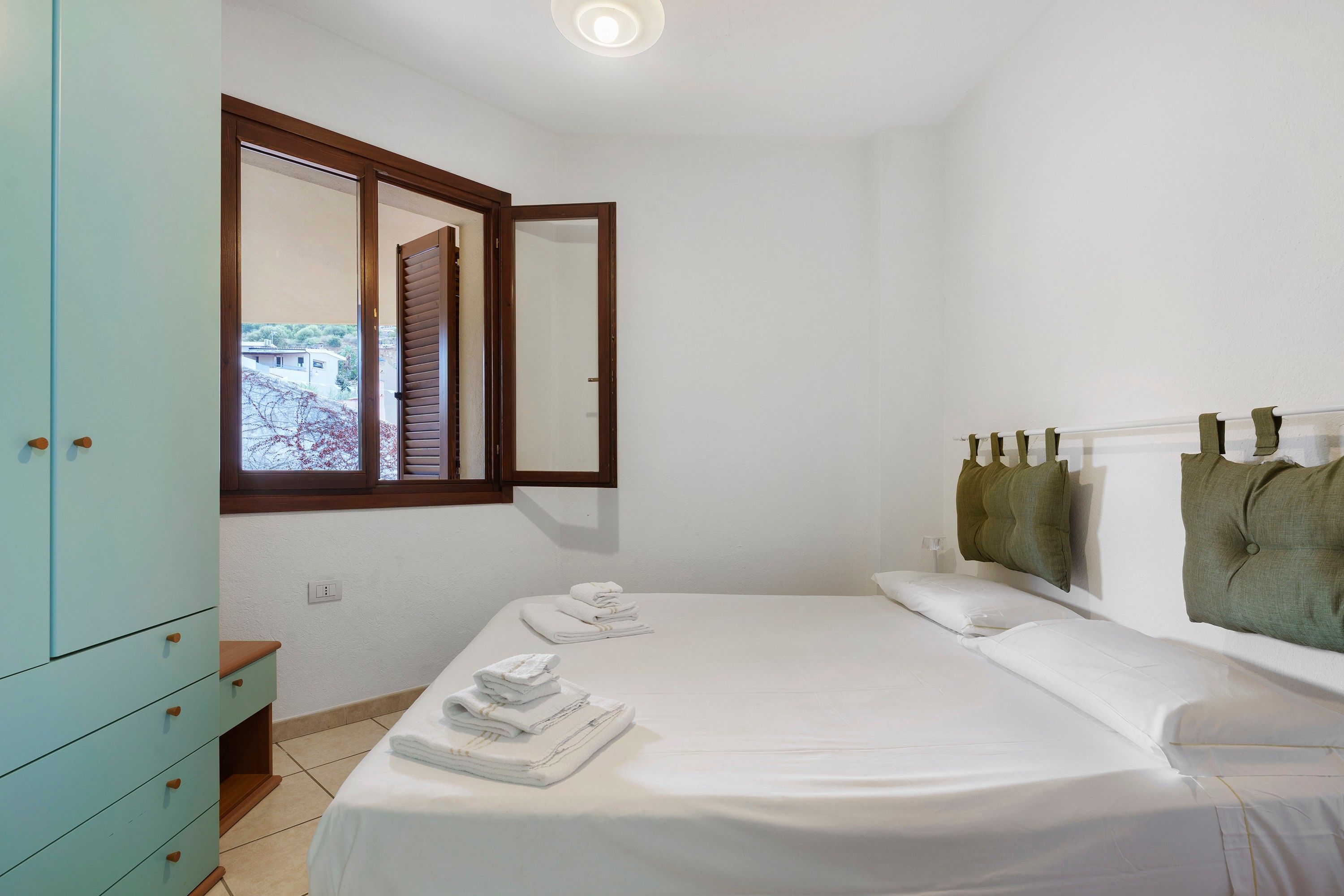 A bedroom with a bed, a wardrobe, a window with blinds, and a view of the outside.