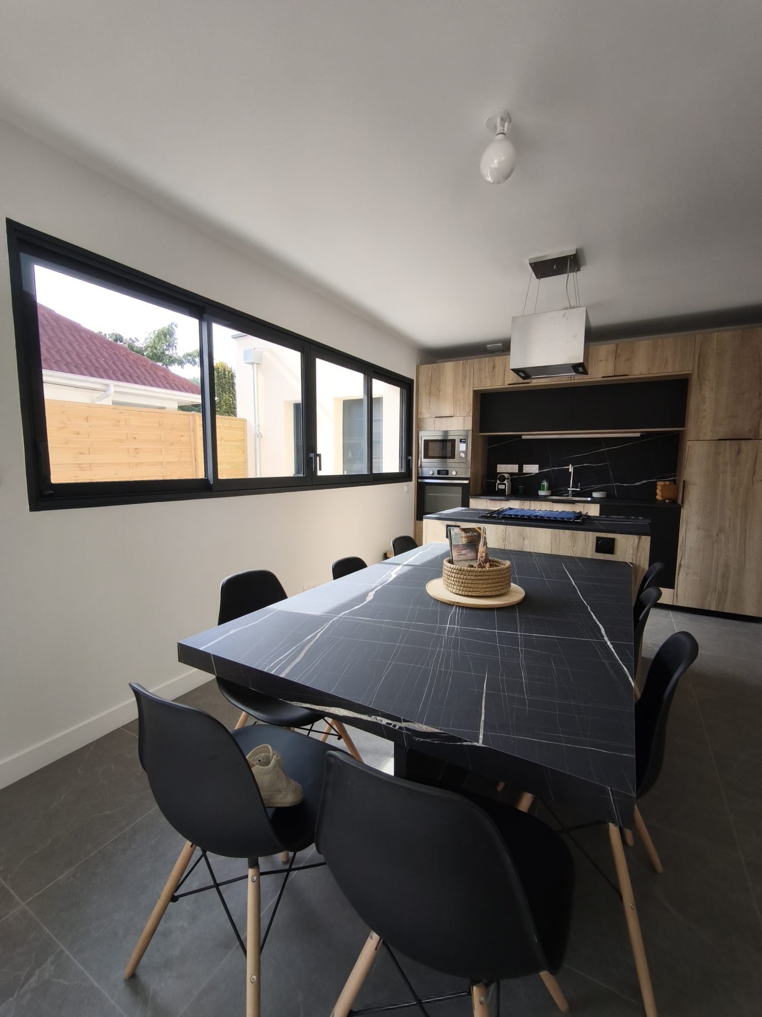 A modern kitchen with a dining area, featuring a large table with chairs, a view through the window, and a built-in oven.