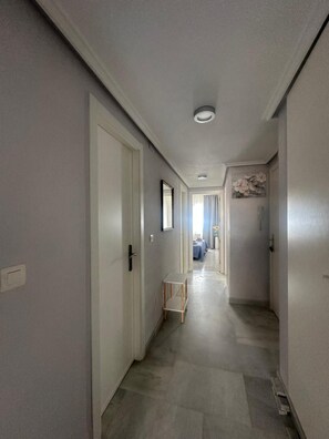 Ein schmaler Korridor mit weißen Wänden und Fliesenboden, der zu einem Raum mit einem Bett und einem Fenster führt.