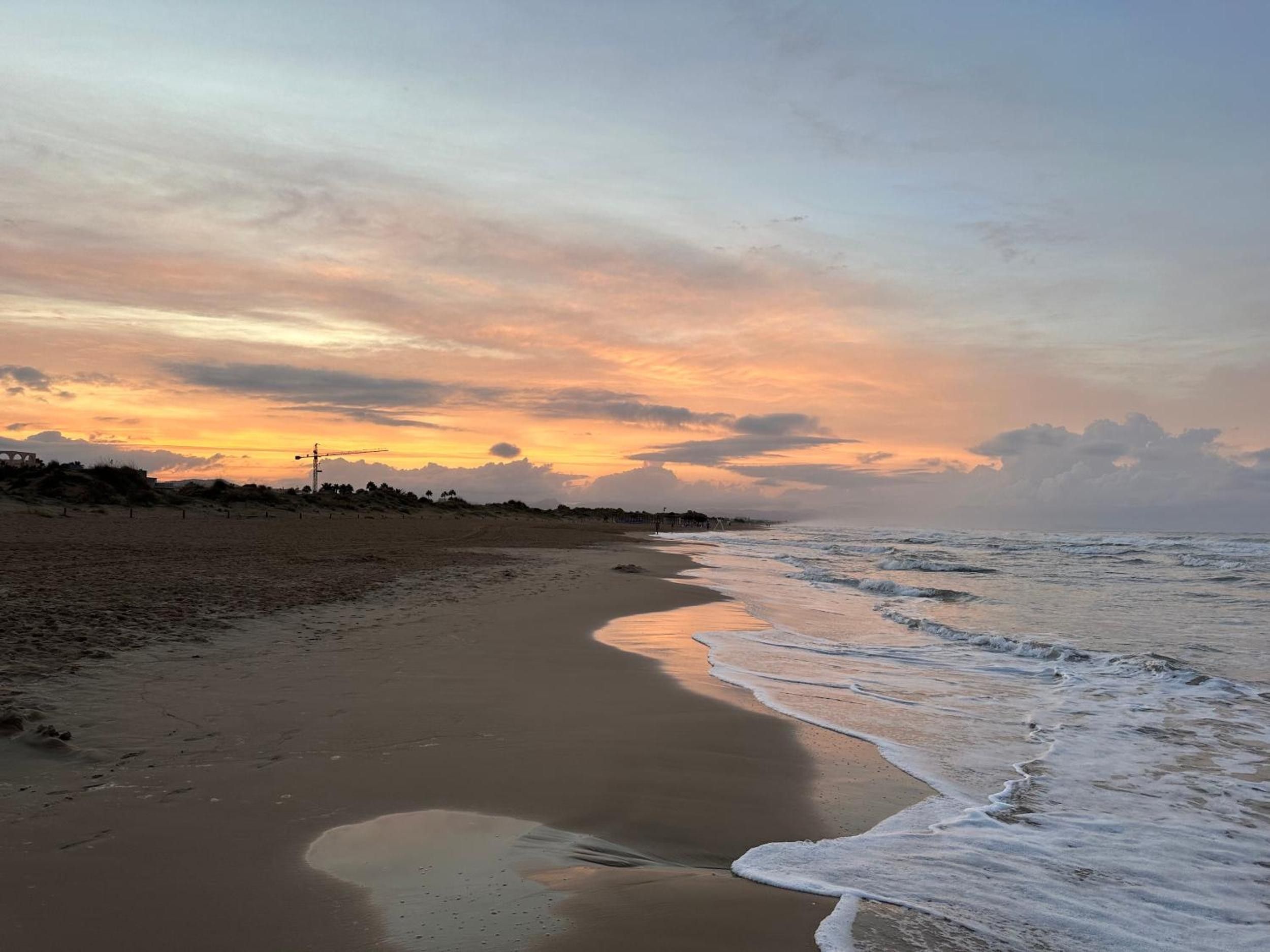 Una playa con una costa curvada, olas acercándose a la orilla y una puesta de sol al fondo.
