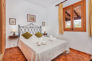 Ein ordentlich bezogenes Bett mit weißen Leinentextilien und zwei Kissen, ein Nachttisch mit einer Lampe, ein gerahmtes Bild an der Wand und ein Fenster mit Vorhängen.
