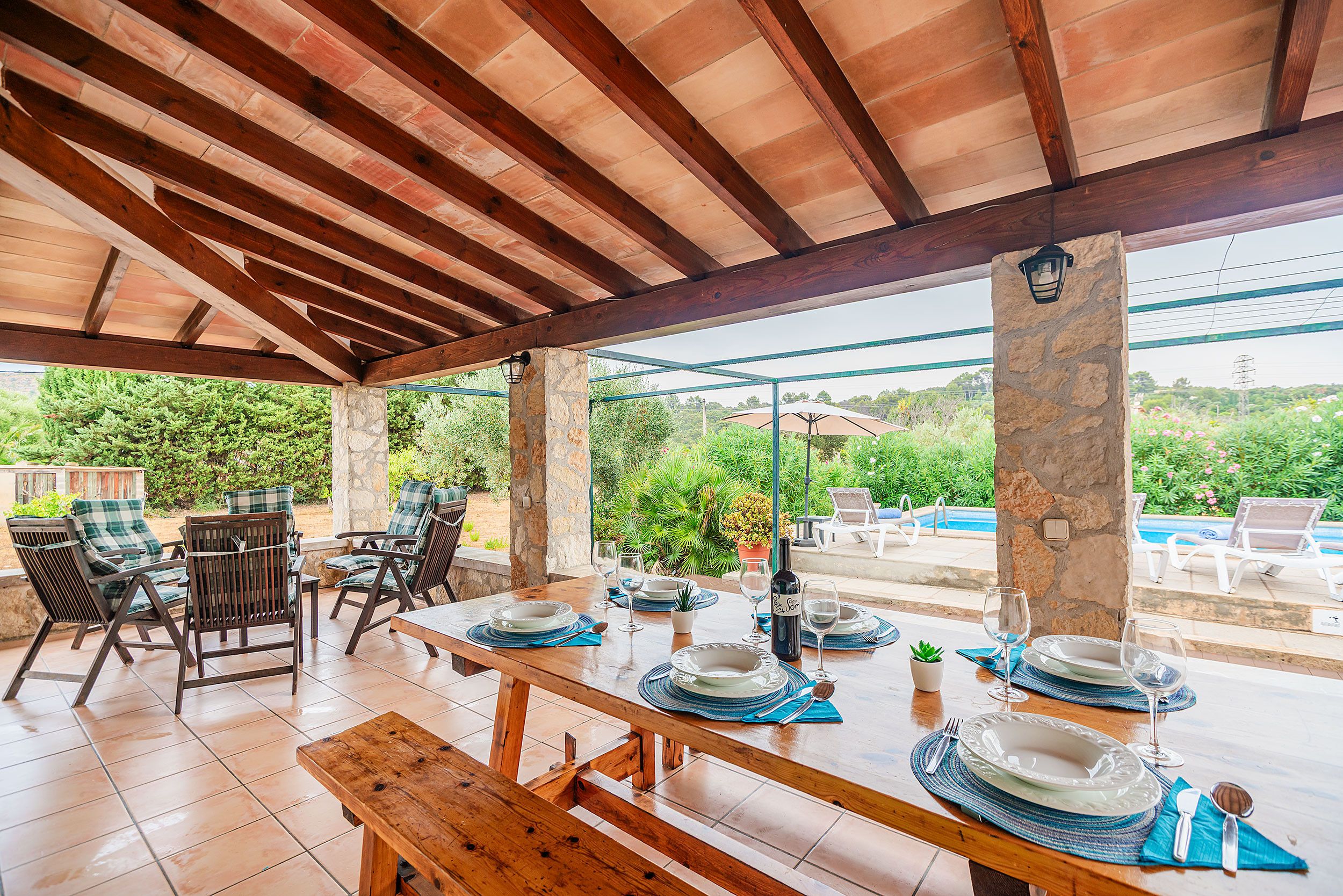 An outdoor patio with wooden tables and chairs, stone pillars, and a view of a swimming pool and greenery.