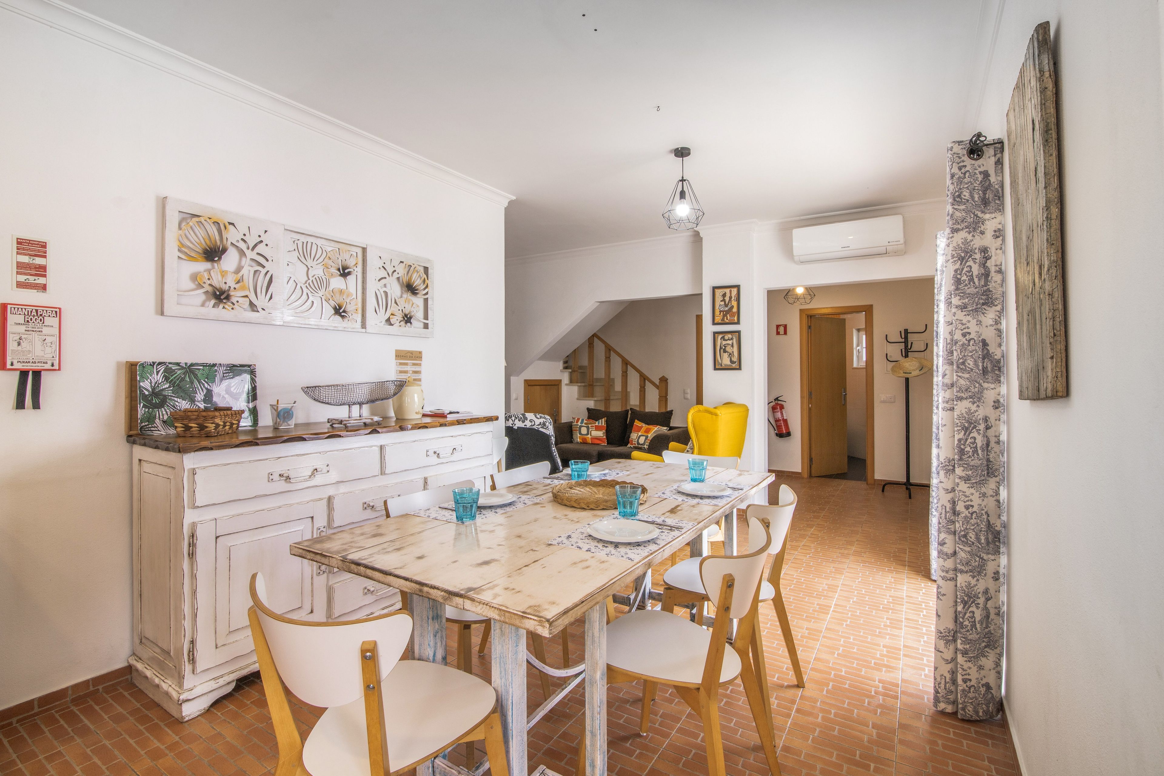 Un comedor con mesa de madera rústica, sillas blancas y una zona de cocina con armarios blancos y una lámpara colgante.