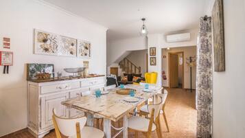 Un comedor con mesa de madera rústica, sillas blancas y una zona de cocina con armarios blancos y una lámpara colgante.