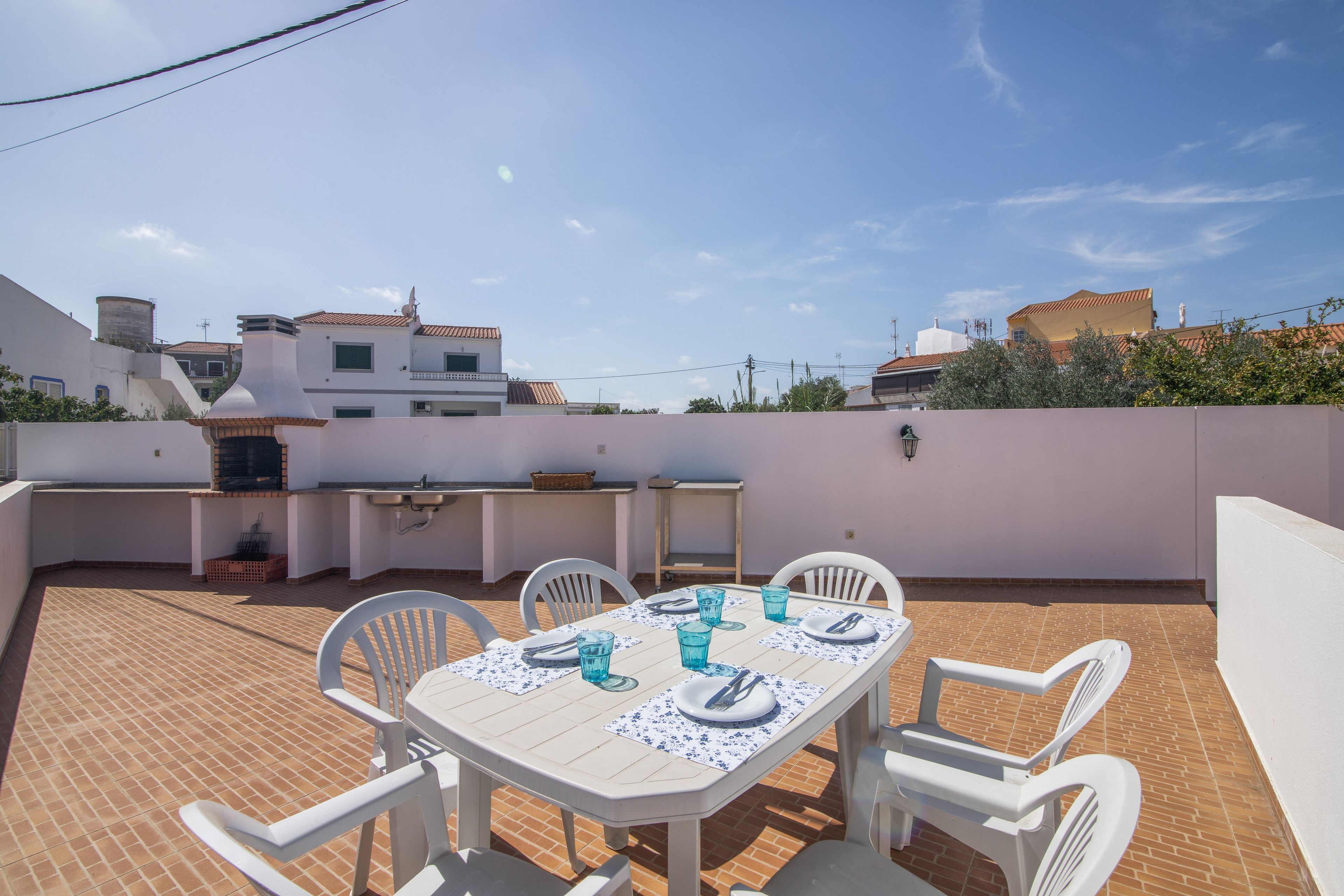 Un patio exterior con una mesa preparada para cuatro personas, rodeado de sillas blancas, bajo un cielo despejado.