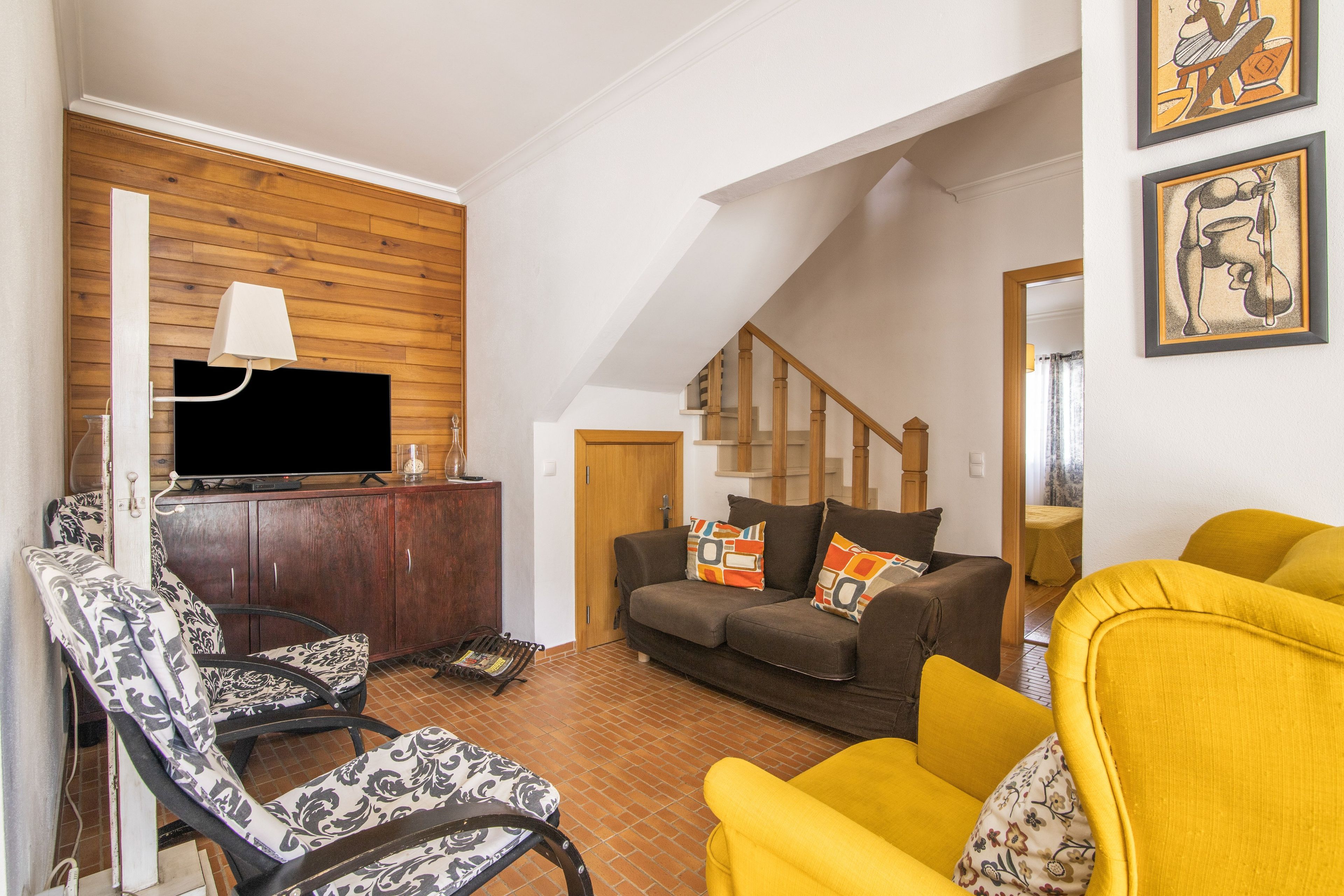 Ein Wohnzimmer mit Fernseher, gemusterten Stühlen, einer Couch mit bunten Kissen und einem gelben Sessel sowie einer Treppe.