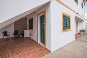 A building with a sloped roof, a door, and two windows with shutters. There is a brick wall, a tiled floor, and a small outdoor seating area with two chairs.