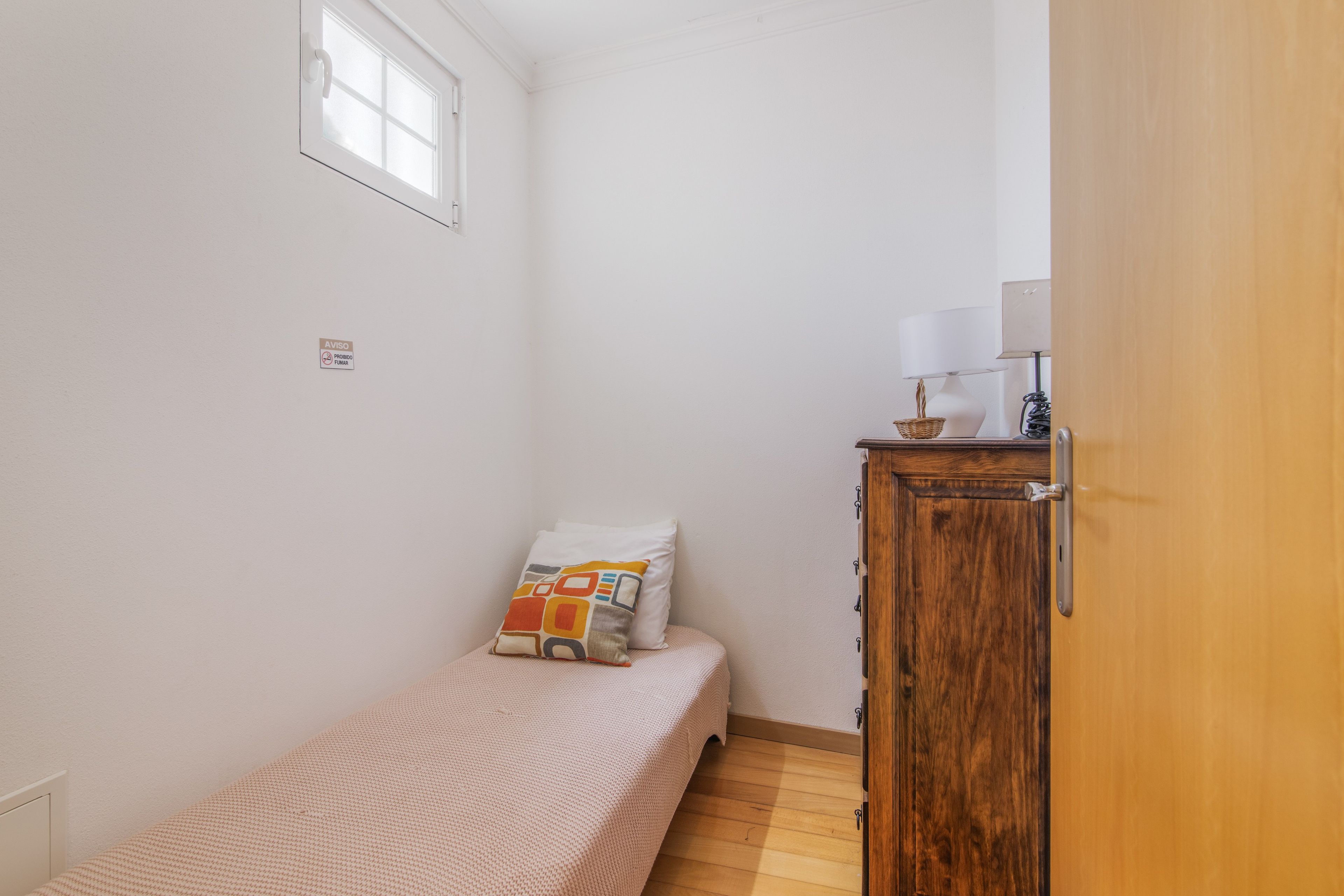 Une petite chambre avec un lit simple, une armoire en bois, une fenêtre et un luminaire fixé au mur.