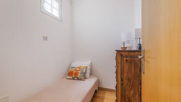 A small, single-bed room with a wooden wardrobe, a window, and a wall-mounted light fixture.