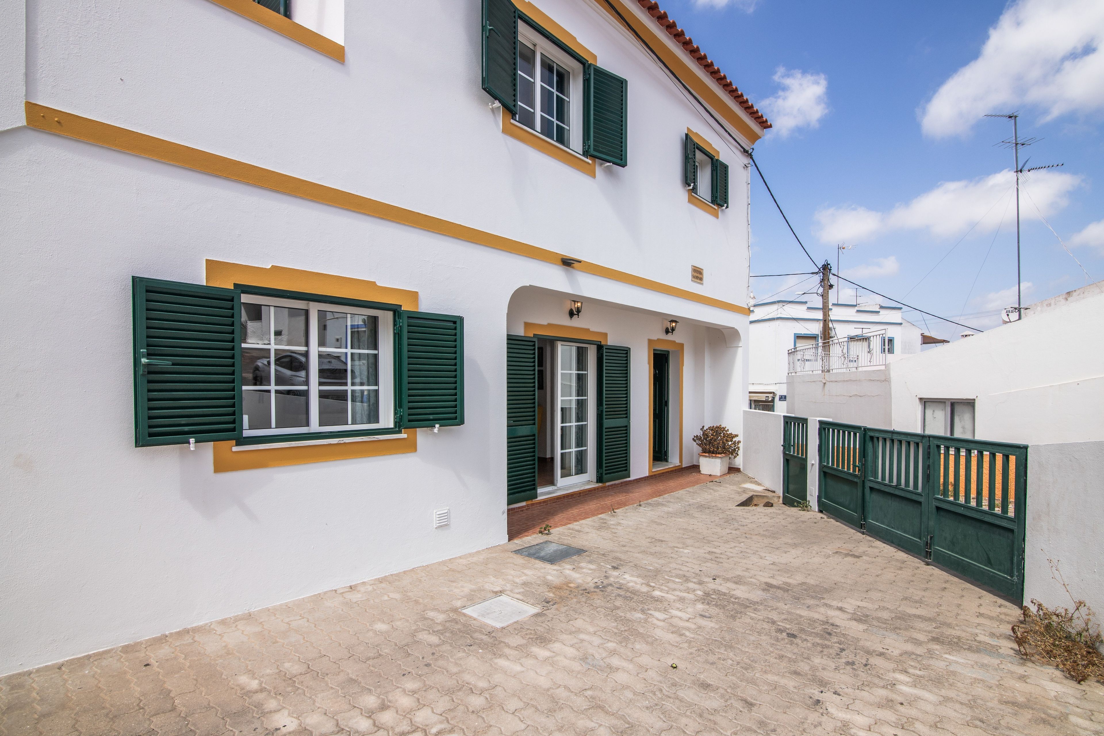 Un edificio de dos plantas con contraventanas verdes, un patio adoquinado y una pared blanca con detalles amarillos.