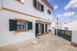 A two-story building with green shutters, a tiled courtyard, and a white wall with yellow trim.